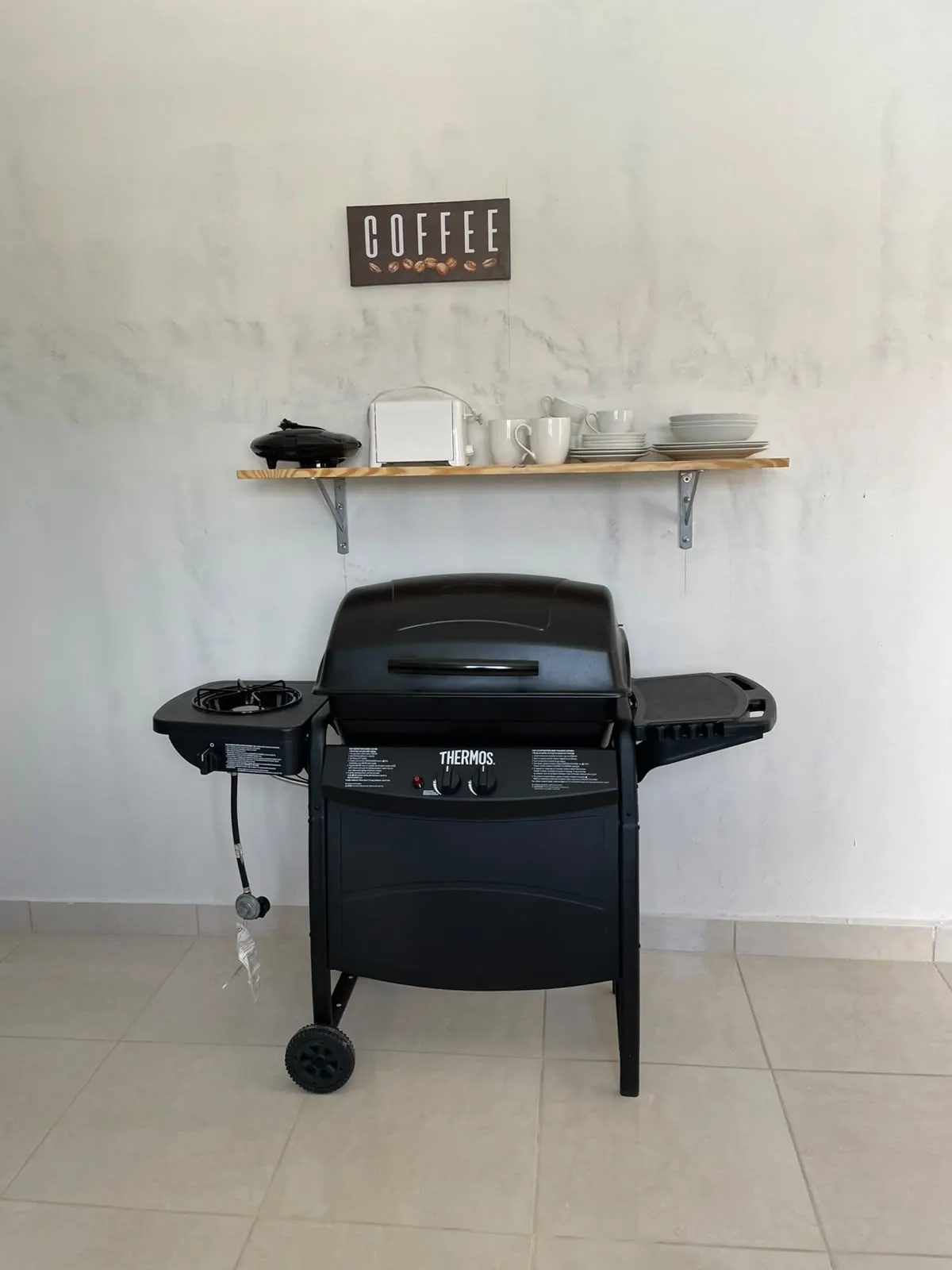 Parrilla a gas junto a un estante de madera con tostadora, tazas y vajilla.